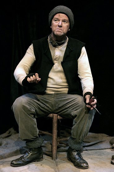 Aidan as Tom Crean performing on stage. He is wearing worn clothes, and wool hat, fingerless gloves and scarf, and holds a pipe, sitting from a stall.